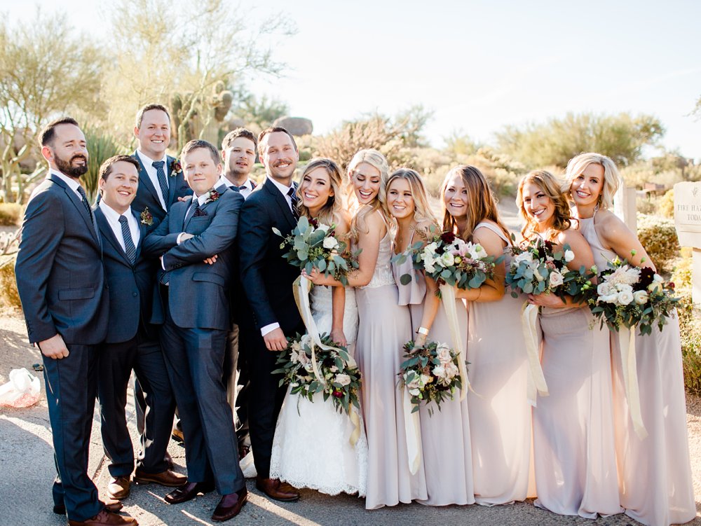 5 bridesmaids in long lilac maxi dresses hugging the bride, female and blonde hair. The bride is wearing a white strapless wedding gown. The groom and groomsmen are on the left side wearing dark blue suits. The scenery is the Arizona desert in Troon North Golf Course.