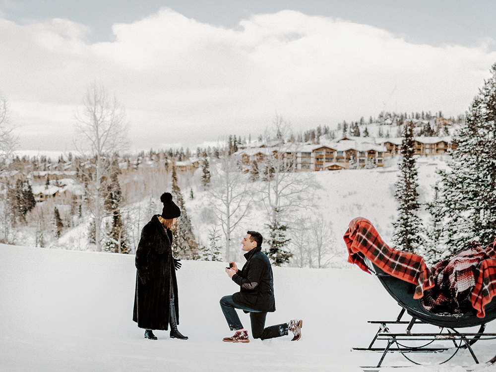 Ali is down on one knee proposing to Sally. The scenic background is in beautiful Deer Valley in Utah. It is filled with white snow and beautiful blue skies.  