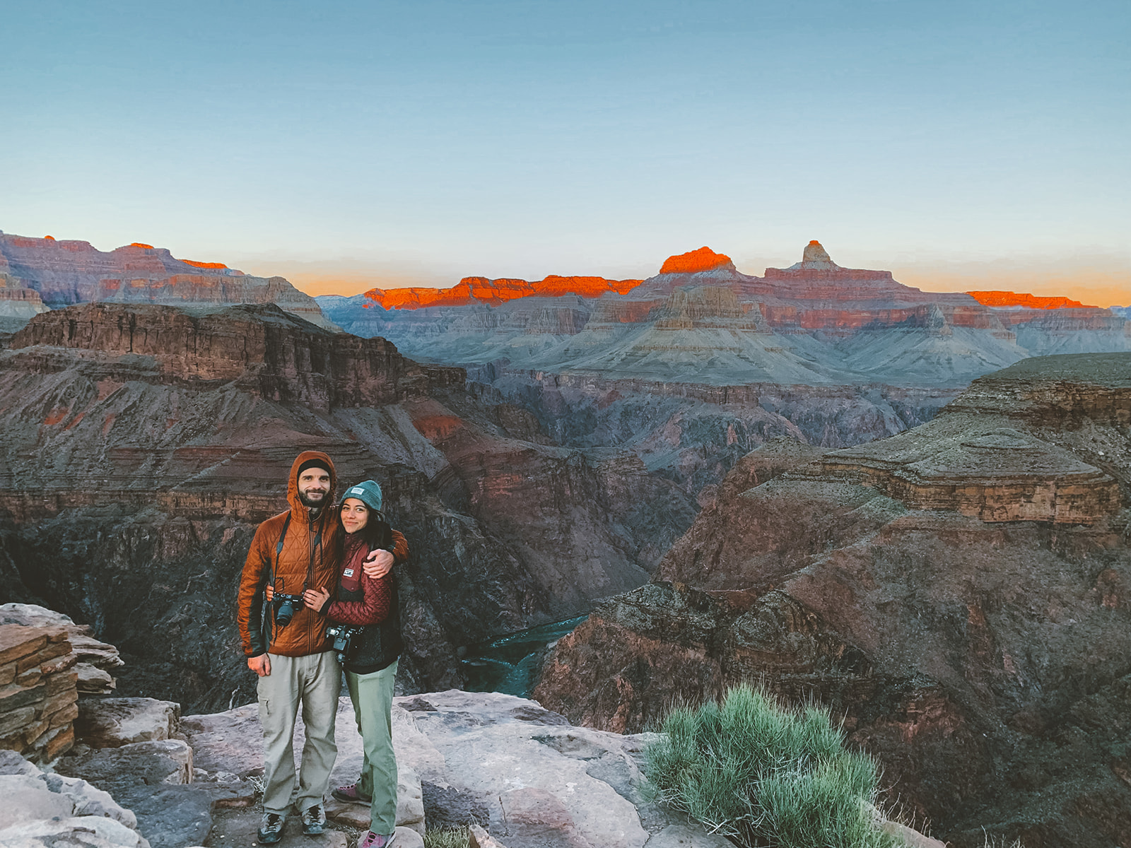 A girl and guy posing, arms wrapped around one another, smiling for the camera. The background is a scenic view of the Grand Canyon