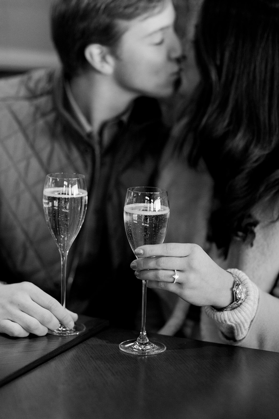 The couple toasting a glass of champagne inside the Stein Eriksen Lodge, Deer Valley. The image is in black and white and you can see both the woman and man holding a glass of bubble in each hand. The woman who was recently proposed to shows off her engagement ring.