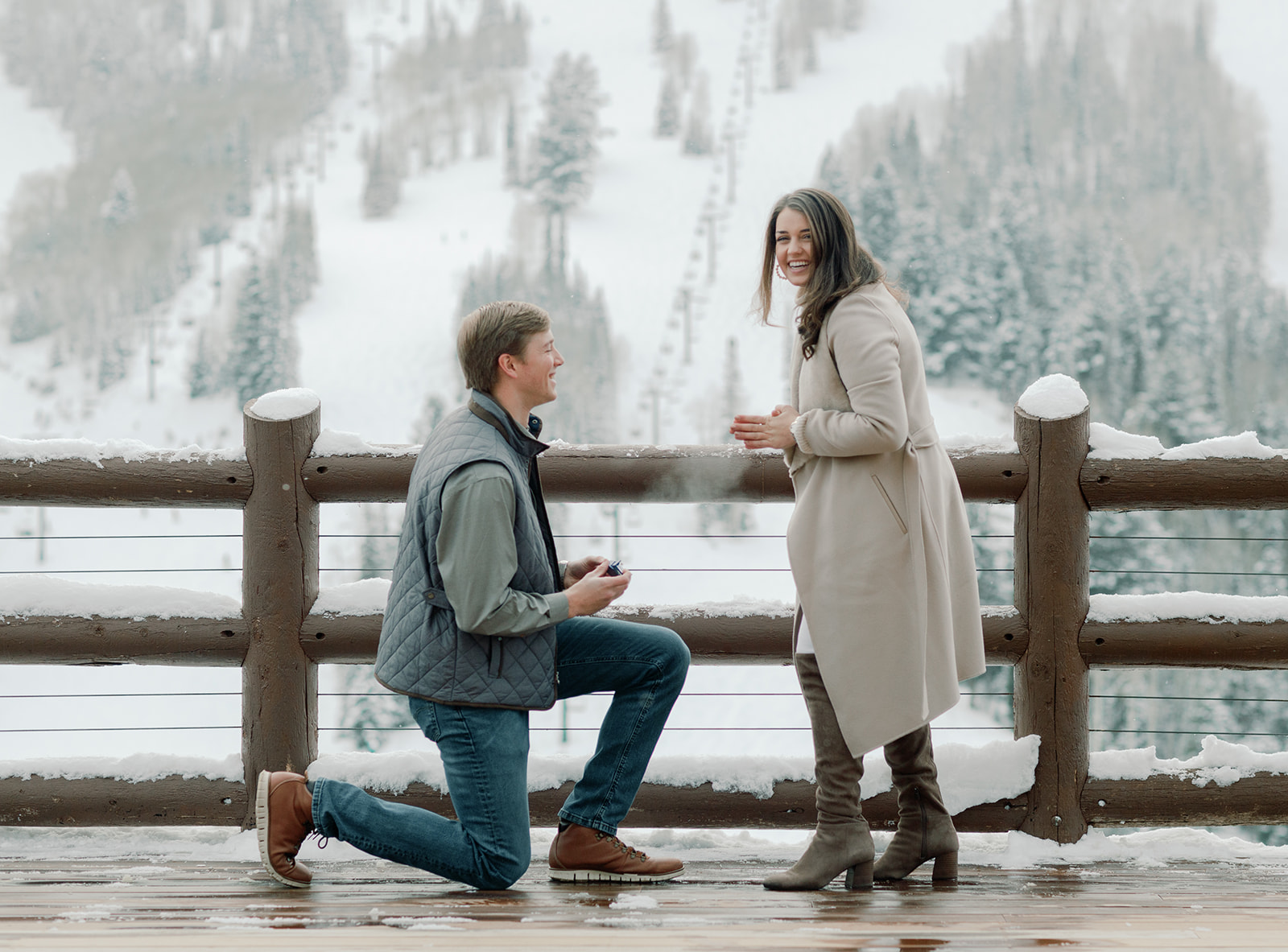  A white male down one knee getting with a ring in his hand. The male has dirty blonde hair and is looking up at his soon to be fiance. He is down on knee proposing to the brunette female. The female is wearing a tan peacoat and is looking up at the camera in complete surprise on her marriage proposal. The scenery in the background are beautiful snowy mountains. The deck they are on is called the "Flagstaff Mountain Deck" and is located at Stein Eriksen Lodge, Deer Valley. 
