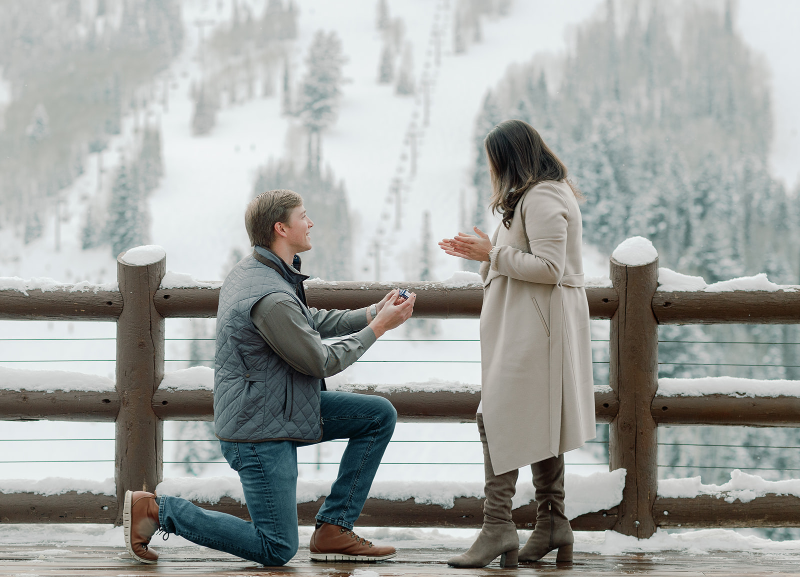 A white male down one knee getting with a ring in his hand. The male has dirty blonde hair and is looking up at his soon to be fiance. He is down on knee proposing to the brunette female. The female is wearing a tan peacoat and is looking down with her hands out in full surprise on her marriage proposal. The scenery in the background are beautiful snowy mountains. The couple had a beautiful proposal at Stein Eriksen Lodge, Deer Valley. 