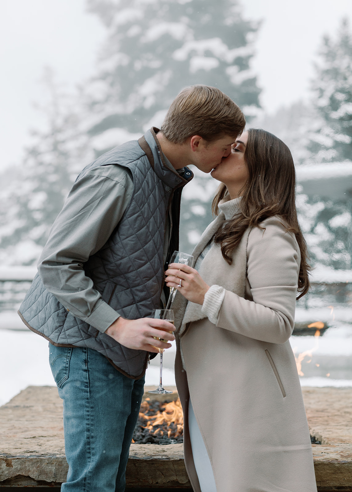 A newly engaged couple sharing a kiss. They each hold a glass of champagne in their hand to cheers and celebrate a marriage proposal. The woman wearing her engagement ring she got from her male fiance. She also wears white pans, a long cream peacoat and a cream sweater turtleneck. The male wears jeans, a nice shirt, with a puffer vest over the button up shirt. 