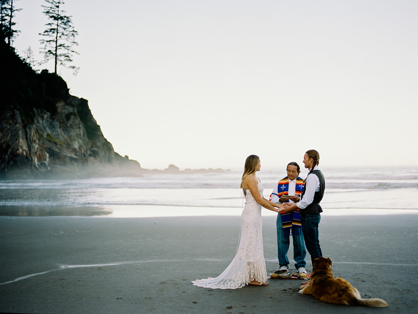 A white female with blonde hair wearing a white boho lace wedding gown. She is holding hands with a male, the groom, and he is wearing blue jeans, a white button up and grey sweater vest. They have their officiant in front of them as they recite their vows for their Oregon Coast Elopement.