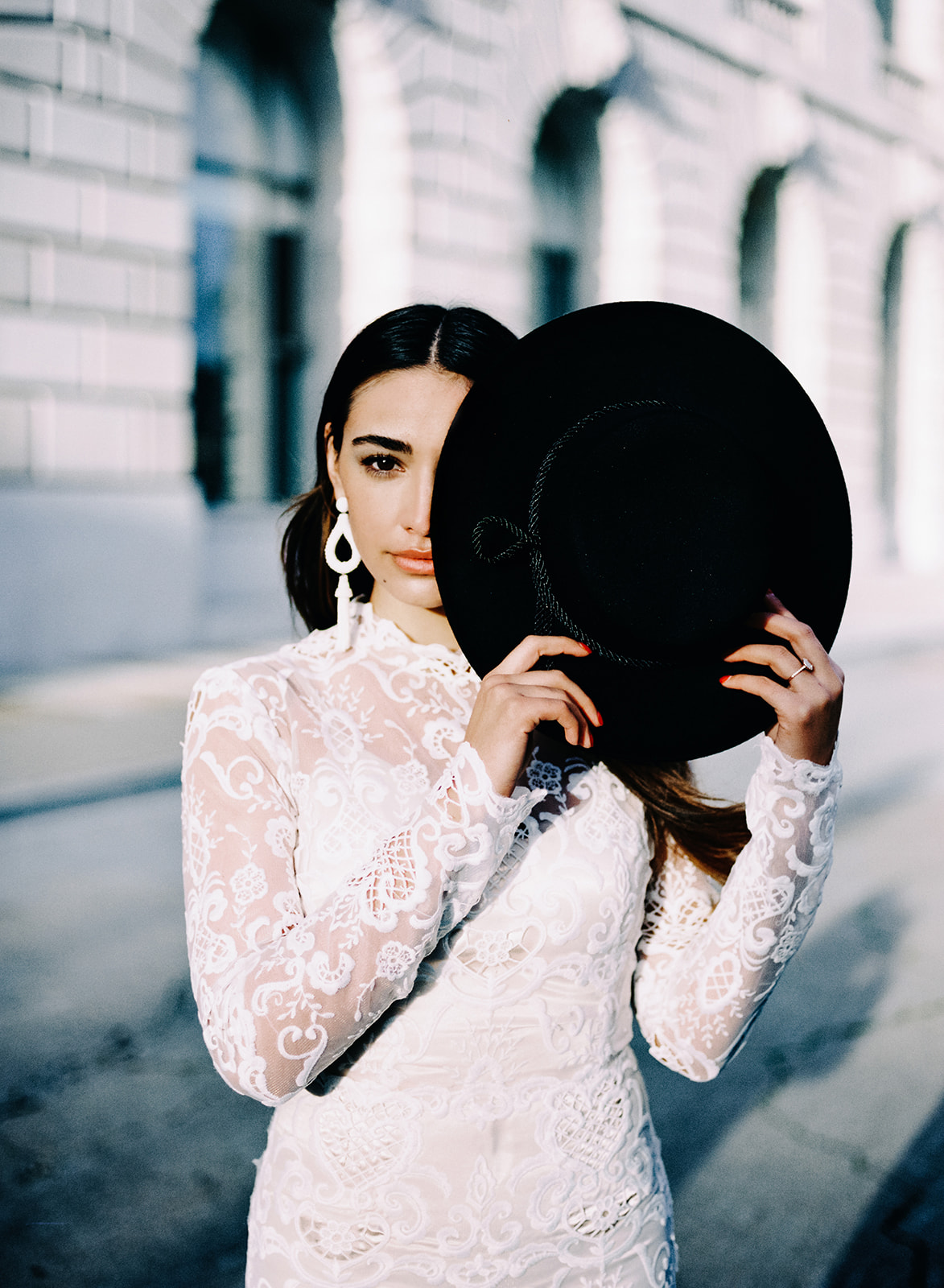 A female with long brunette hair posing for this San Francisco Bridal styled shoot. She holds up a black hat that covers half of her face. She wears a long sleeve lace fitted wedding gown. Her earrings are long and white.