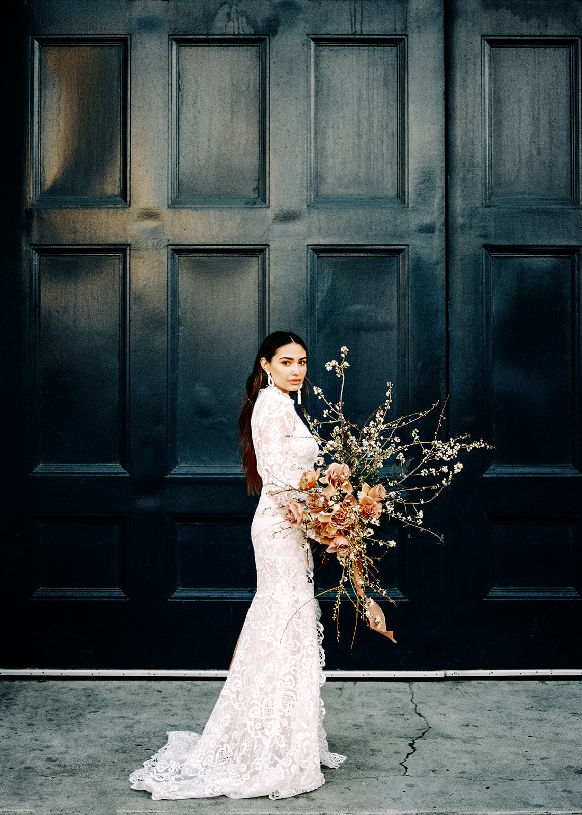 A female with long brunette hair posing for this San Francisco Bridal styled shoot. She holds a bridal bouquet of large florals, long stems and pink roses. She wears a long sleeve lace fitted wedding gown. Her earrings are long and white. The background of the door is black and cement floor is dark grey.