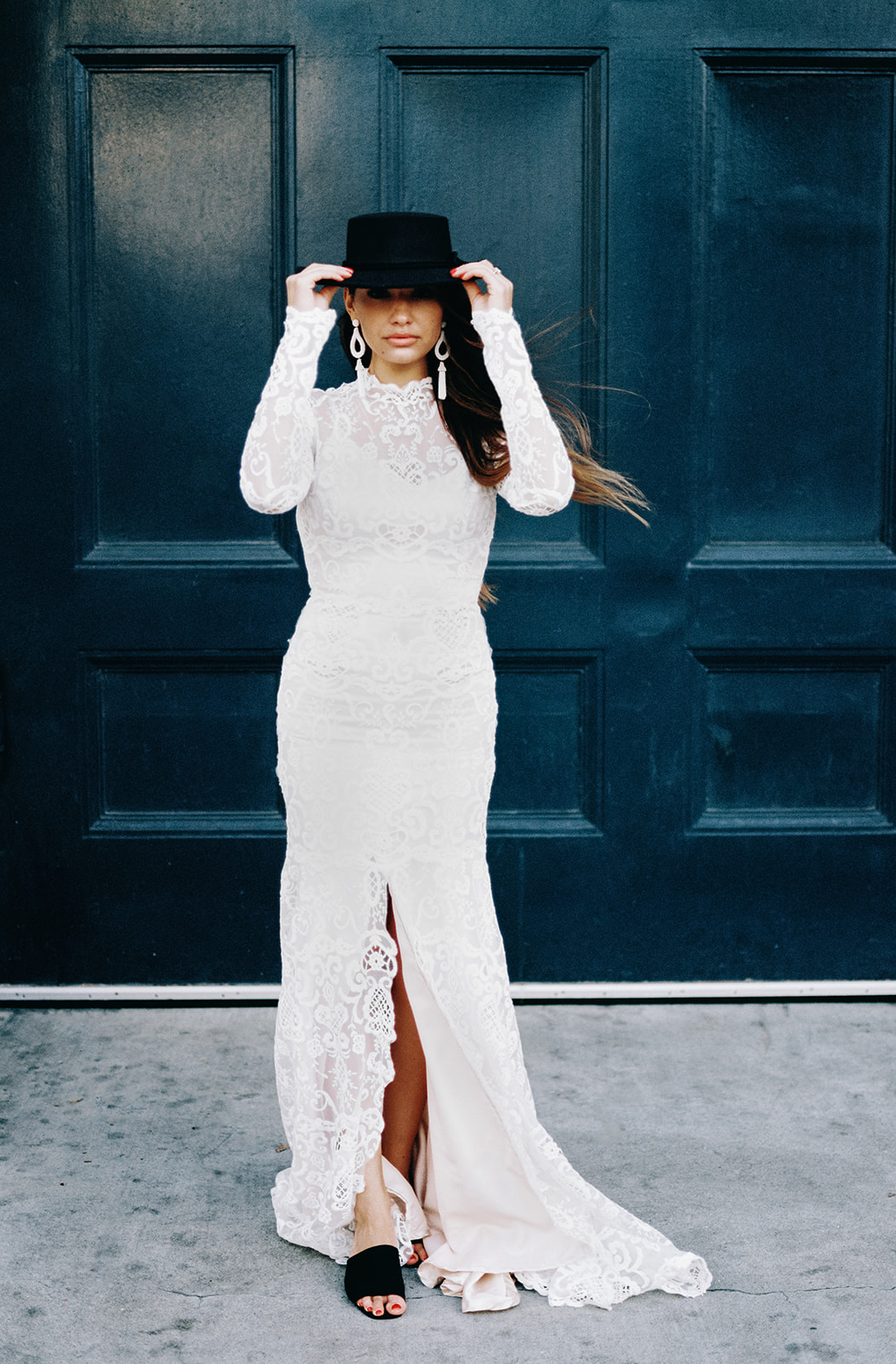 A female with long brunette hair posing for this San Francisco Bridal styled shoot. She holds a black hat that she wears on her head. She wears a long sleeve lace fitted wedding gown. Her earrings are long and white. The color of the door in the background is black.