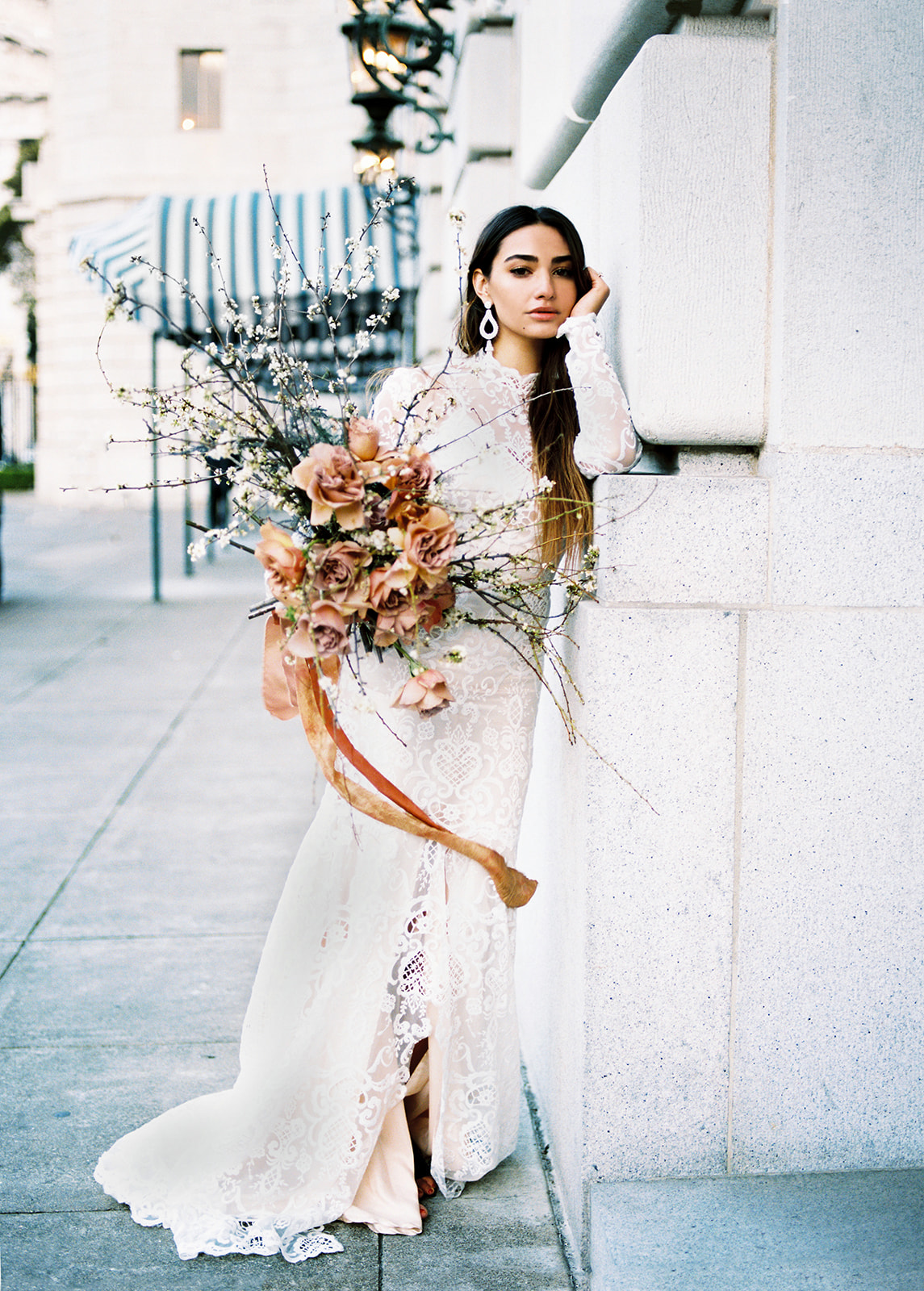 A female with long brunette hair posing for this San Francisco Bridal styled shoot. She holds a bridal bouquet of large florals, long stems and pink roses. She wears a long sleeve lace fitted wedding gown. Her earrings are long and white. She is leaning with one arm up against a light grey cement wall.