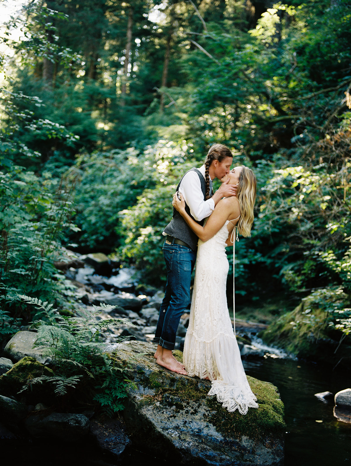 A male, the groom, and a female, the bride, sharing a kiss on top of a grey rock. Behind them is greenery everywhere. There are at the Oregon coast.