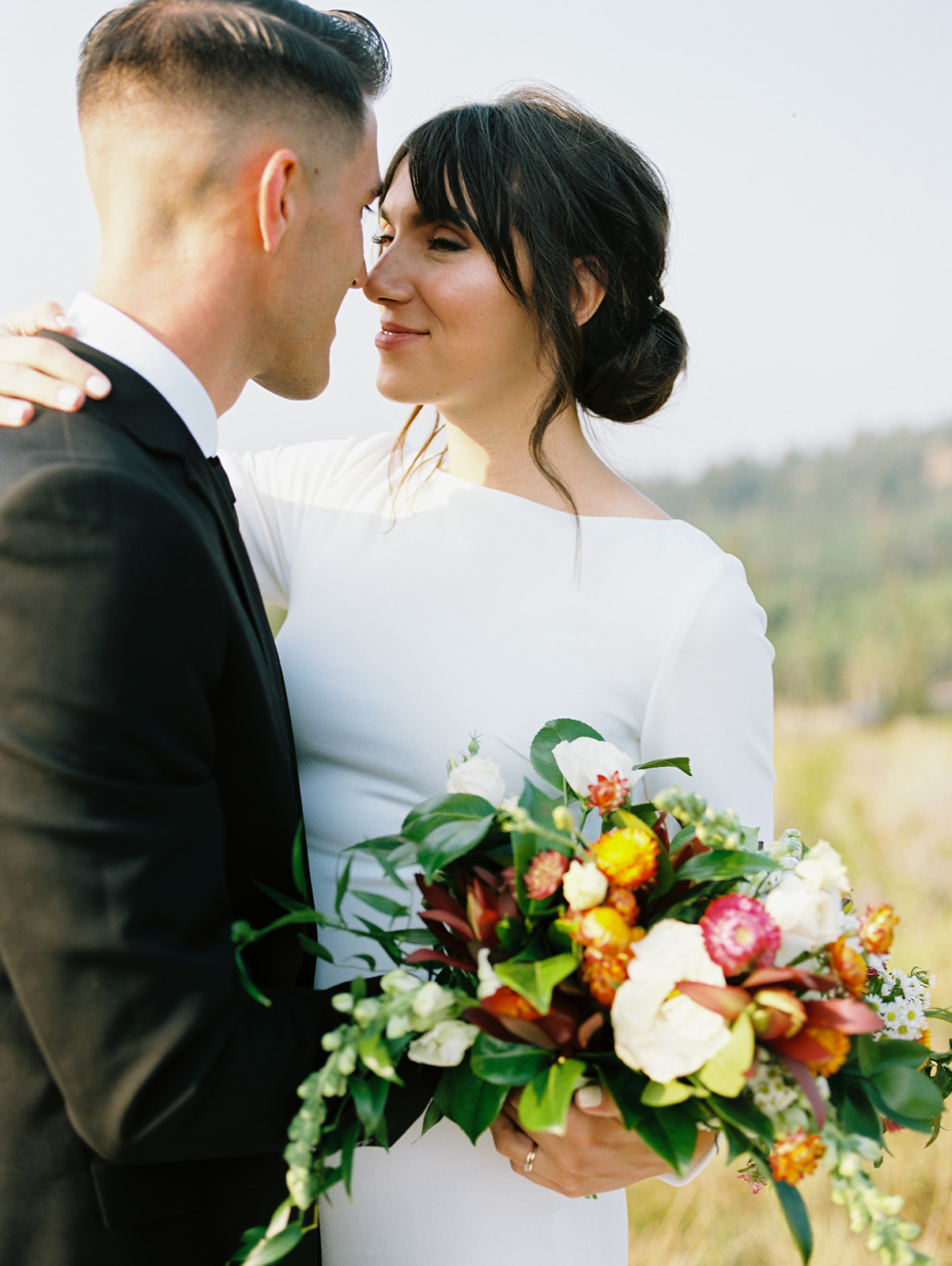 Couple portraits with the bride holding her colorful floral bouquet. She has black hair done in an updo. Wearing a white long sleeve fitted wedding gown.