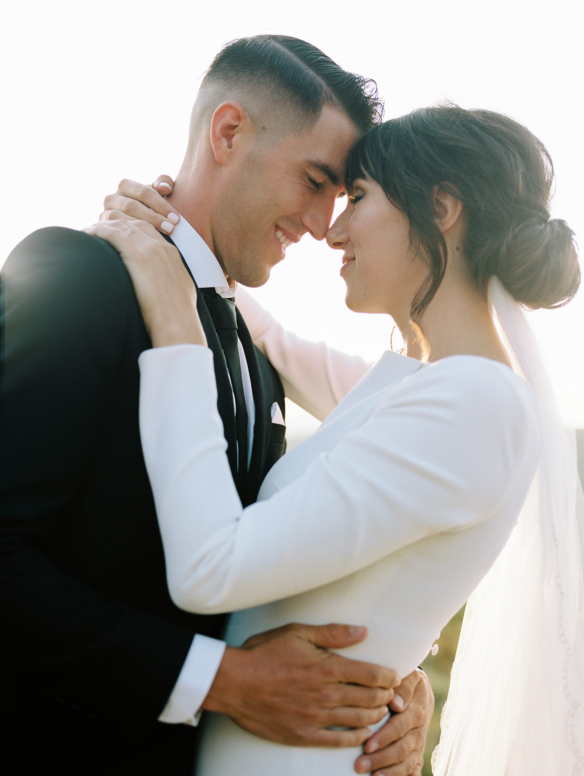 An image of couple portraits. The bride wears a white long sleeve fitted wedding gown. Where the groom wears a black suite. The couple look are facing each other with arms wrapped around one another.