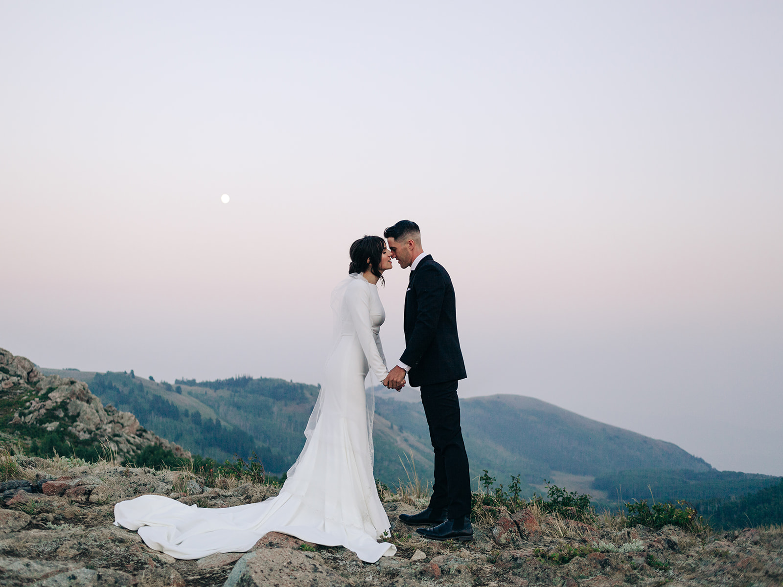 An image of couple portraits with a scenic full moon background. Taken around sunset. The bride wears a white long sleeve fitted wedding gown. Where the groom wears a black suite.