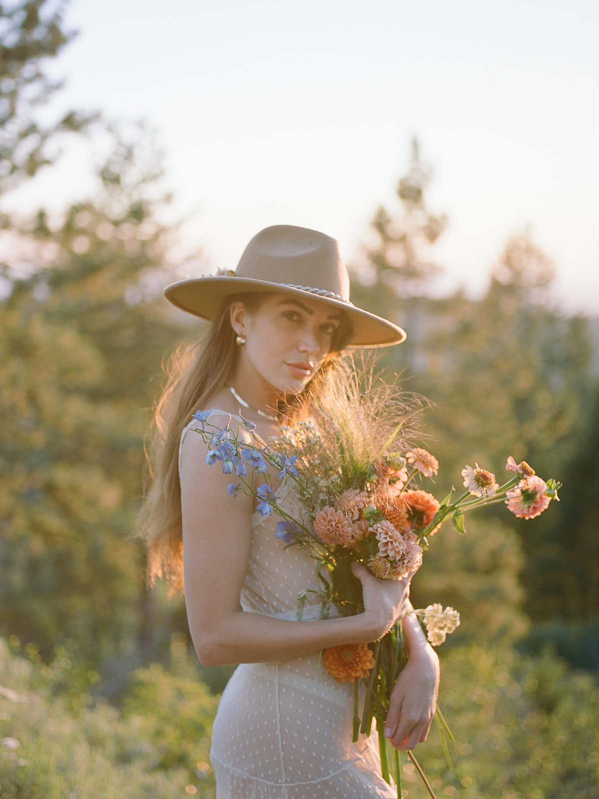 Woman posing for a summer photo shoot. She holds wildflowers and wears a wide brim hat. Backdrop is the hills of the Pacific Northwest. 