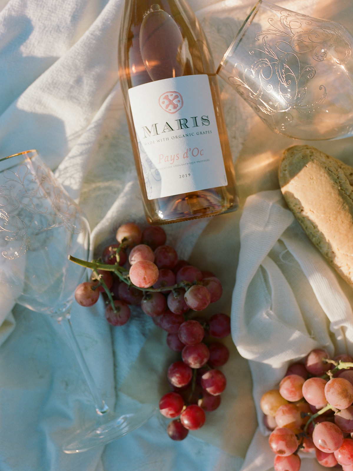 A picnic with wine, wine glasses, grapes, and bread. Set on a flowy white blanket. 