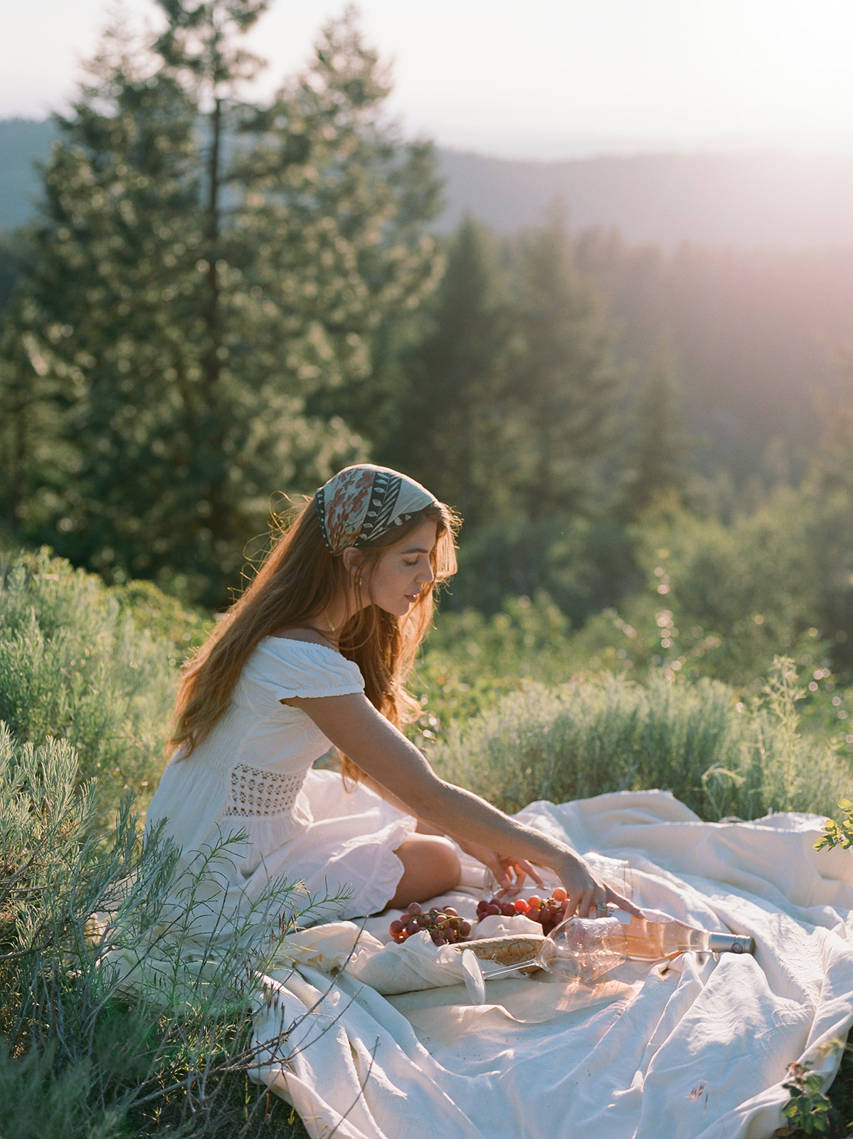 Woman posing for a picnic inspired photoshoot. She poses on a white blanket with wine and grapes 