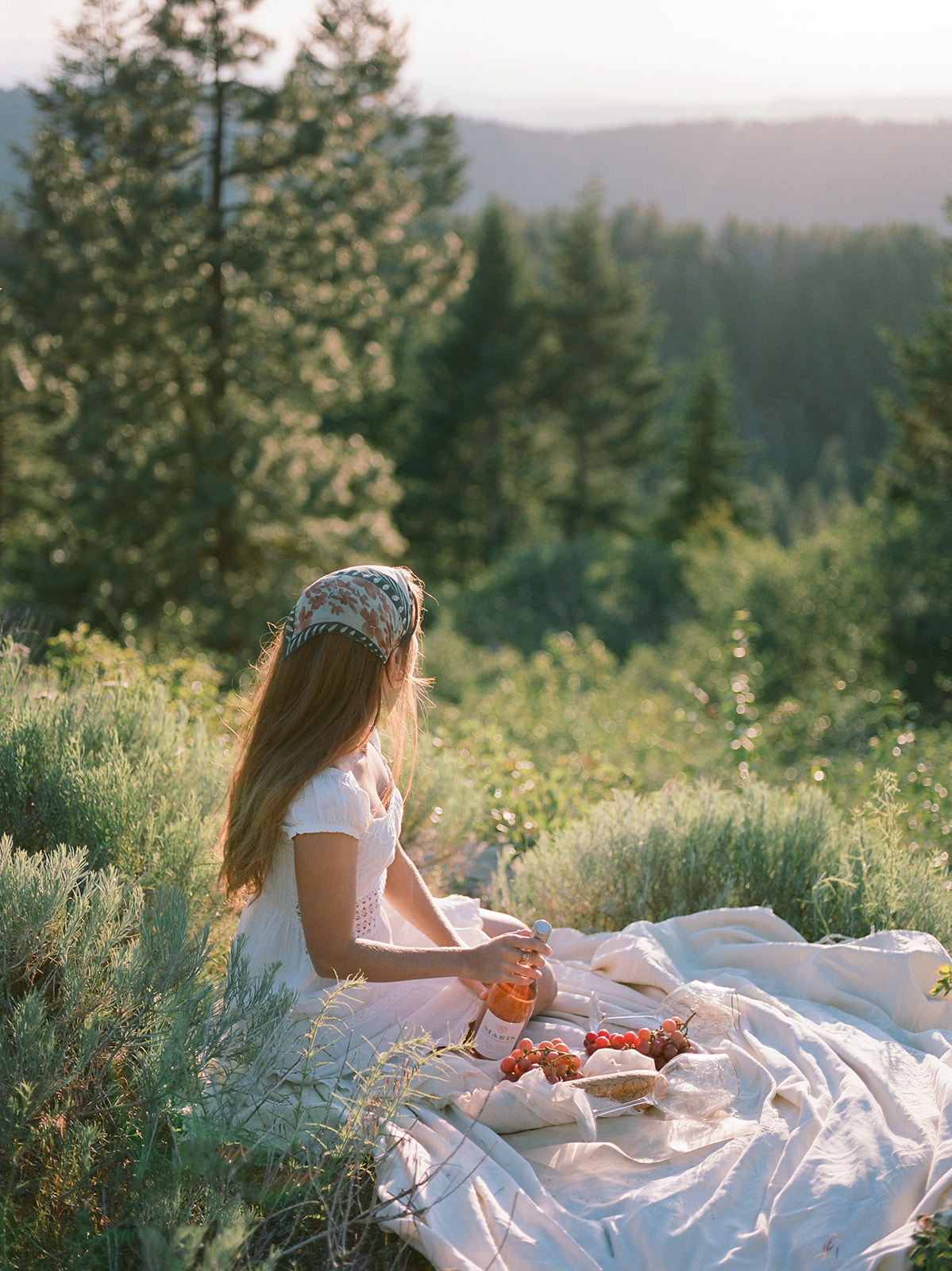 A woman in a flowy white dress and bandana having a picnic in a field. She is in the pacific northwest. 