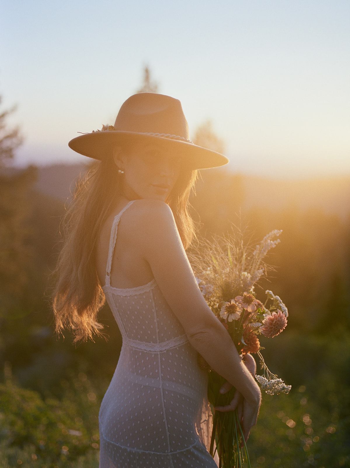A woman poses for a summer photo shoot in the pacific northwest. She holds a bouquet of wild flowers and wears a wide brim hat. 