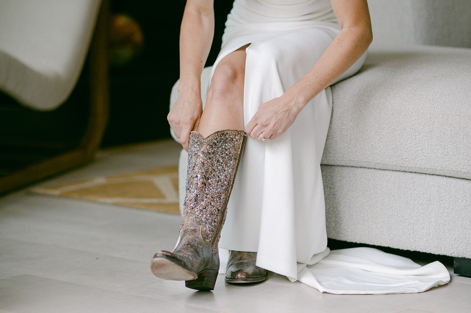 a rustic modern wedding in the high mountains of Utah. The bride is a caucasian female wearing a white crepe fabric wedding dress. She is putting on her sequenced cowboy boot.
