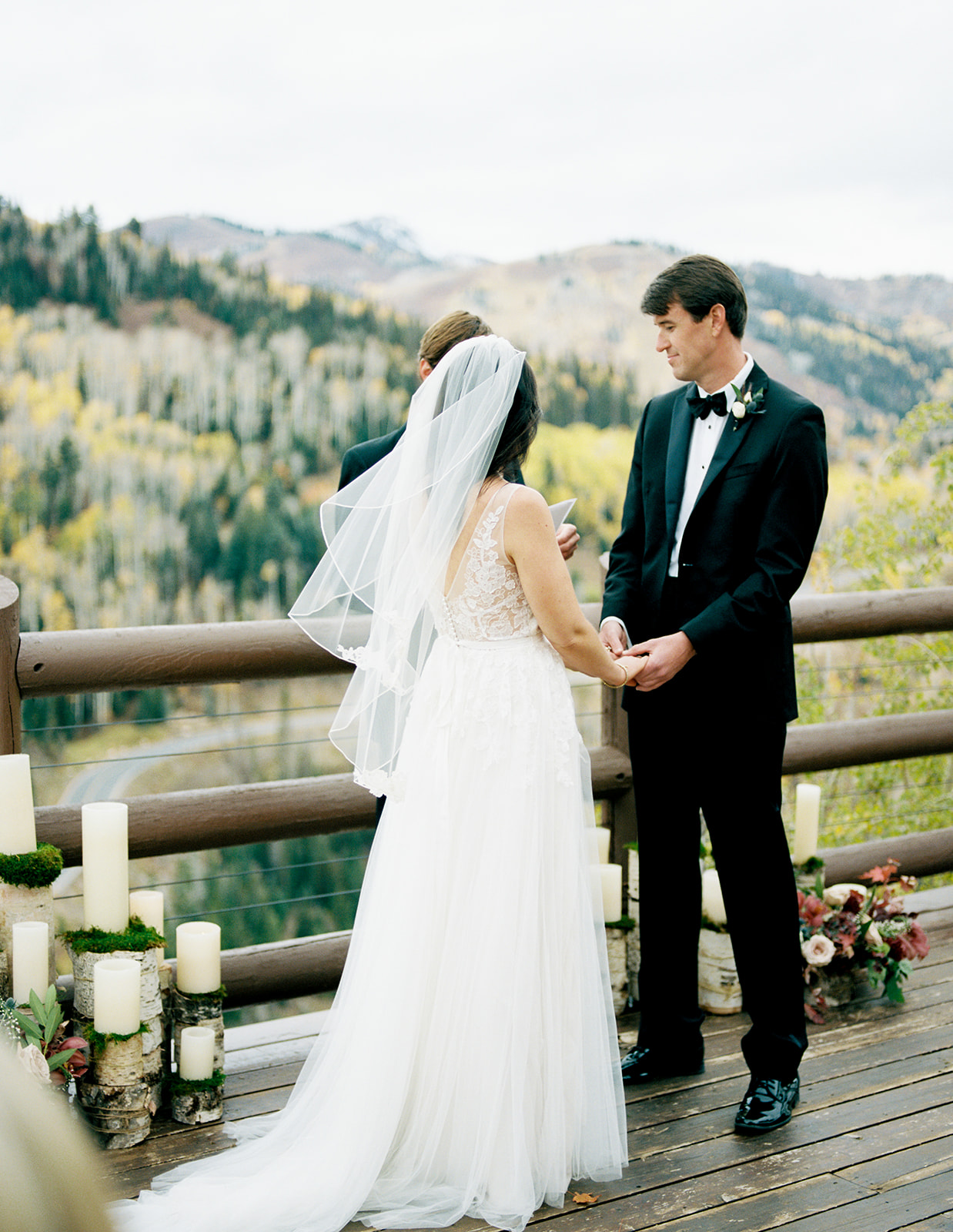 A fall wedding in Park City Utah. The bride is caucasian with black hair wearing a white short veil and white lace gown. The groom is male, caucasian and wearing a black tux with a black bow tie and white under button up shirt. He has dark brown/black hair and is holding the bride's hand during the ceremony.