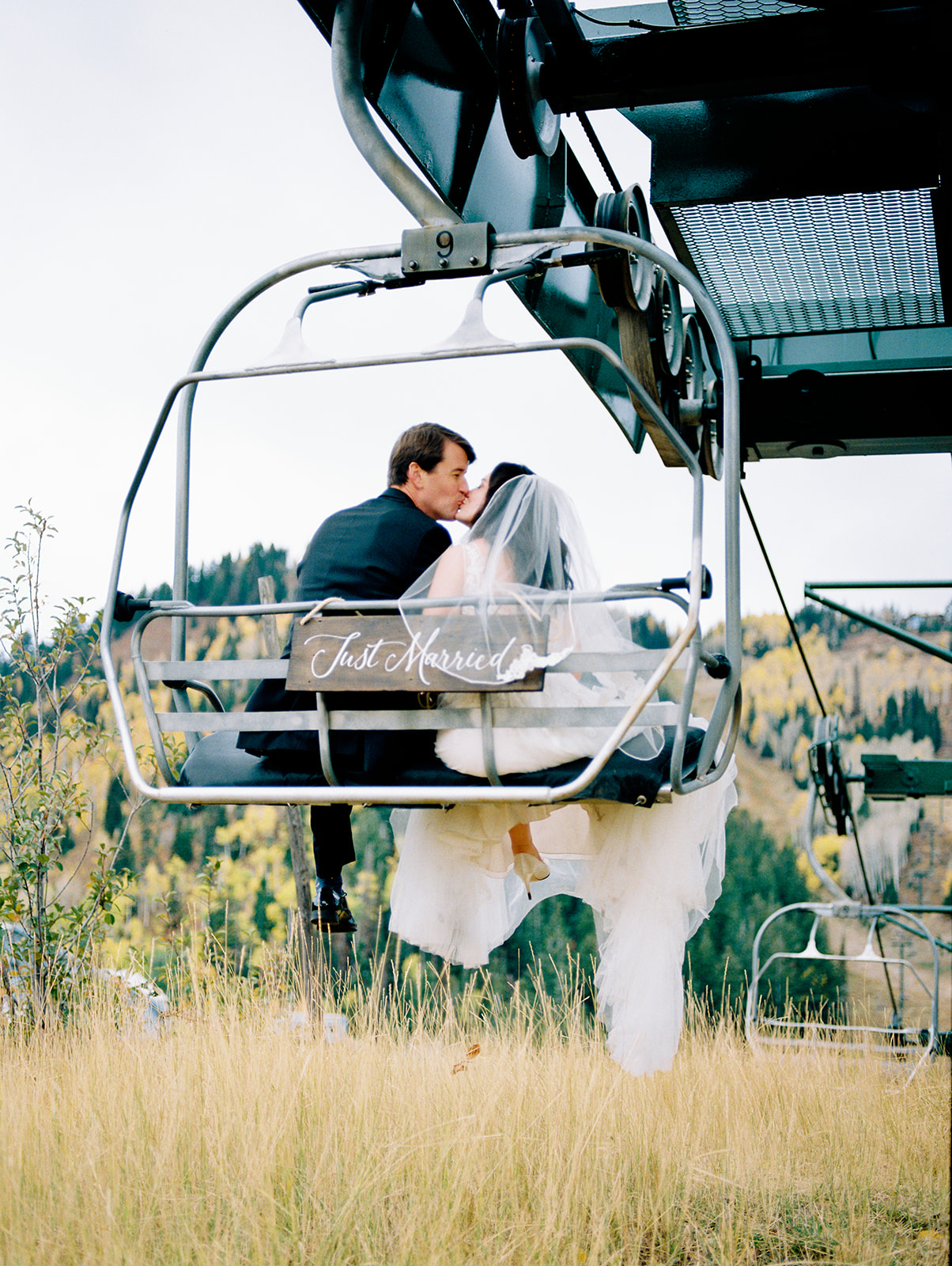 A fall wedding in Park City Utah. The bride wearing a white veil and groom in black tux are sitting on the lift that takes them back to their reception.