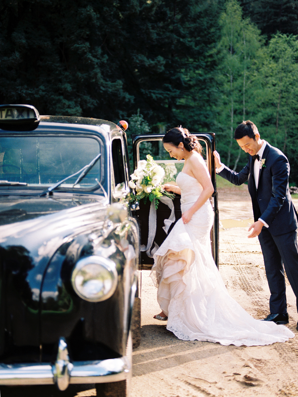 a bride in a white dress and groom in a black suit getting into their black vintage car at their Redwoods wedding venue