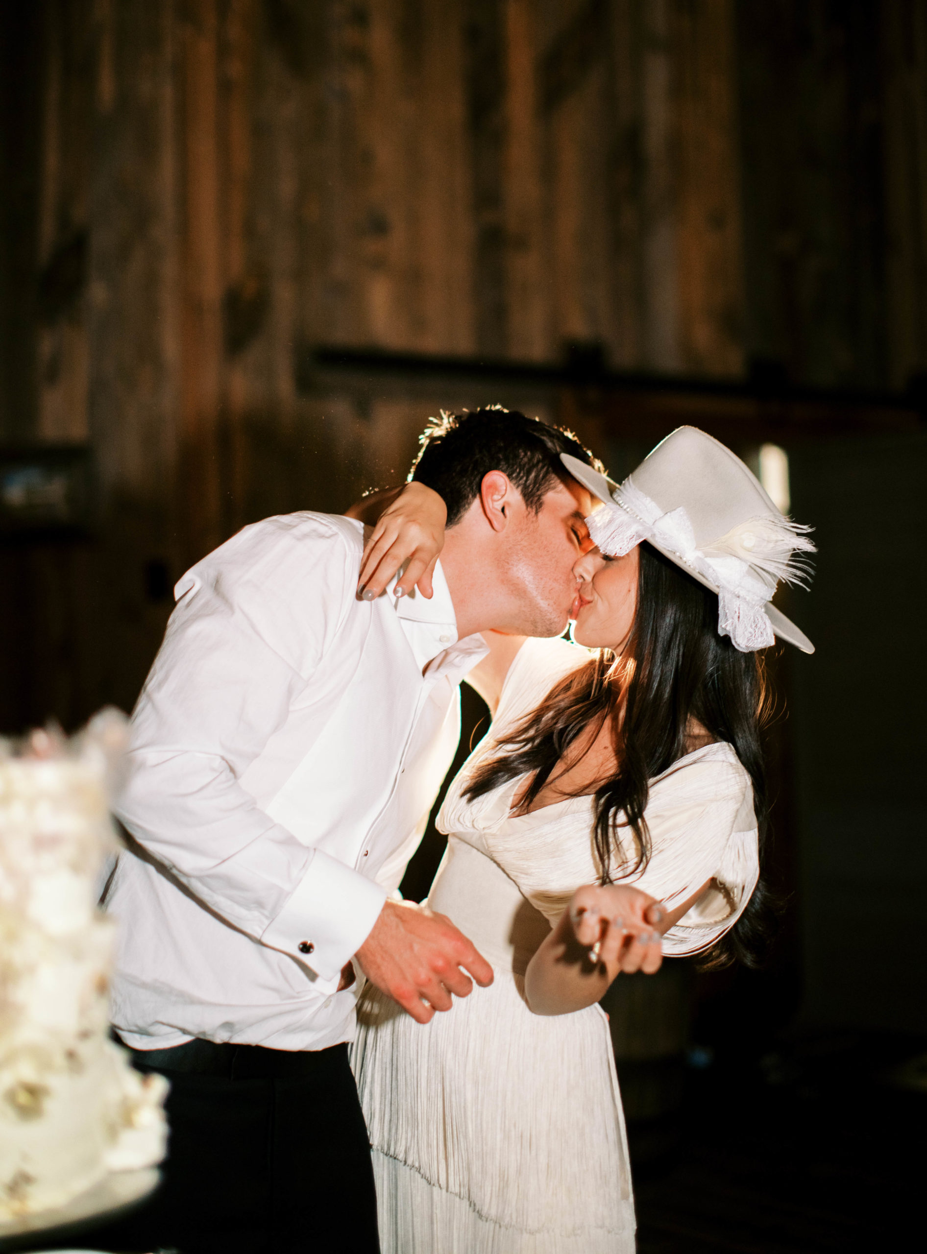 bride and groom kiss while cutting their cake at their utah ranch wedding. the bride wears a vintage wedding reception dress and white bridal hat. photo by megan robinson photography
