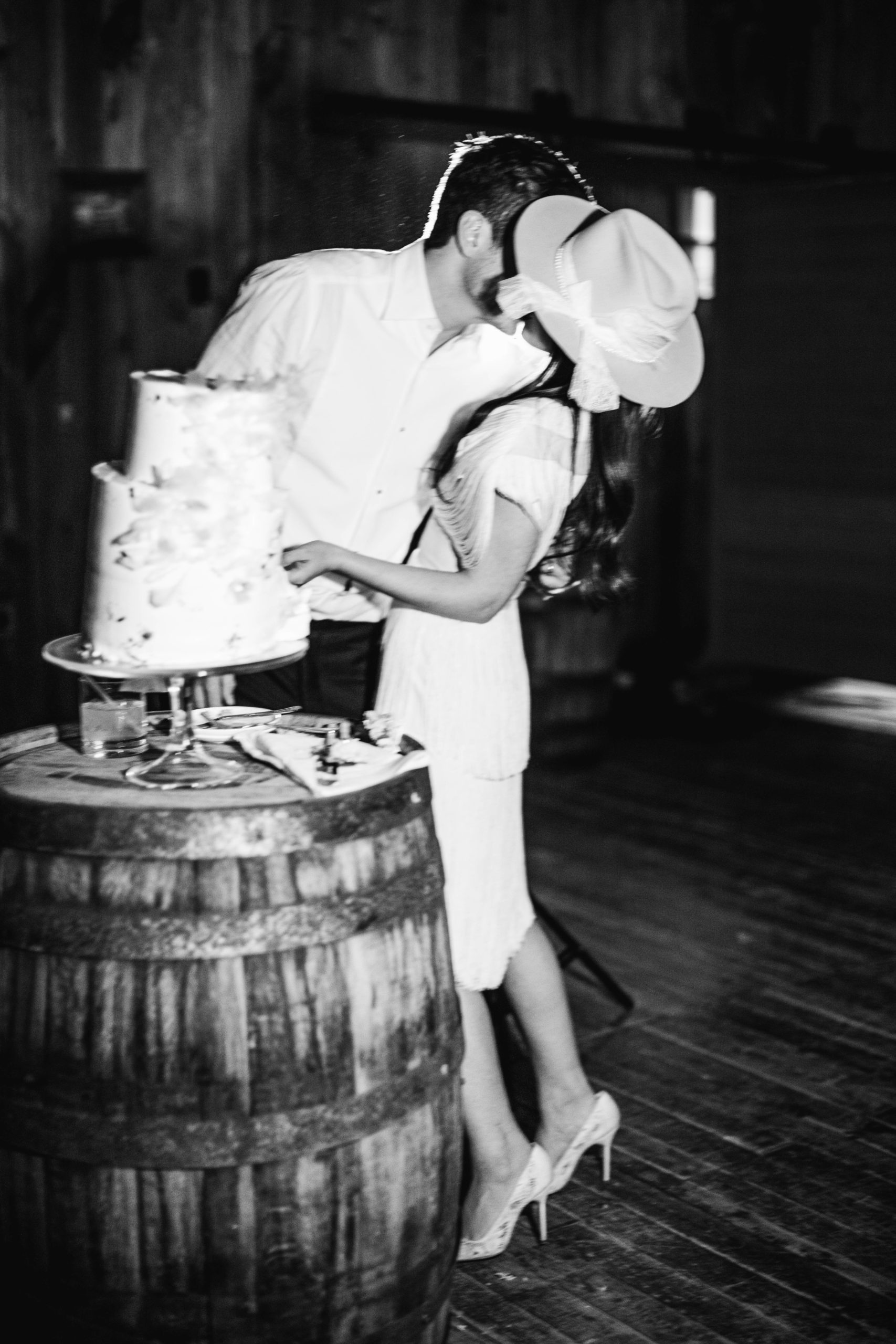 bride and groom kiss while cutting their cake at their utah ranch wedding. the bride wears a vintage wedding reception dress and white bridal hat. photo by megan robinson photography