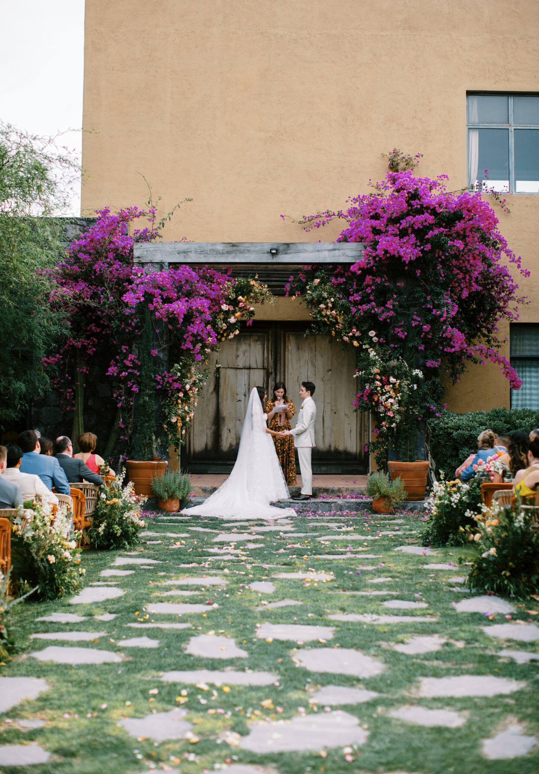 purple lush bougainvillea trees in a green ceremony space in Mexico the bride and groom hold hands as the officiant speaks, and the guests look at them 