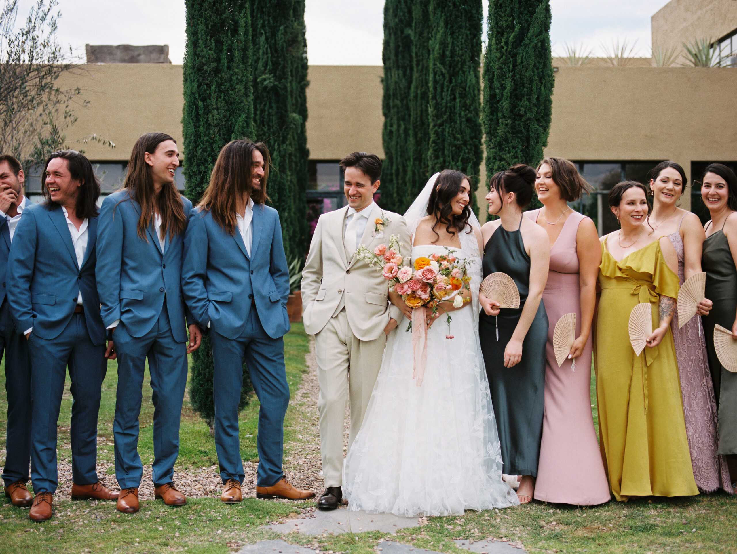 bridal party photos with the bride and groom smiling with groomsmen in blue suits, bridemaids in colorful dresses