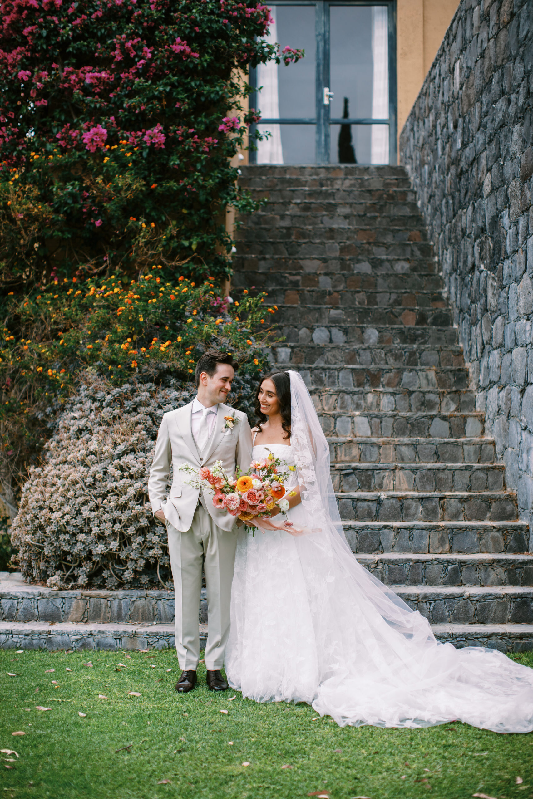 bride and groom smiling at eachother in front of cobblestone stairs and purple lush bougainvillea trees 