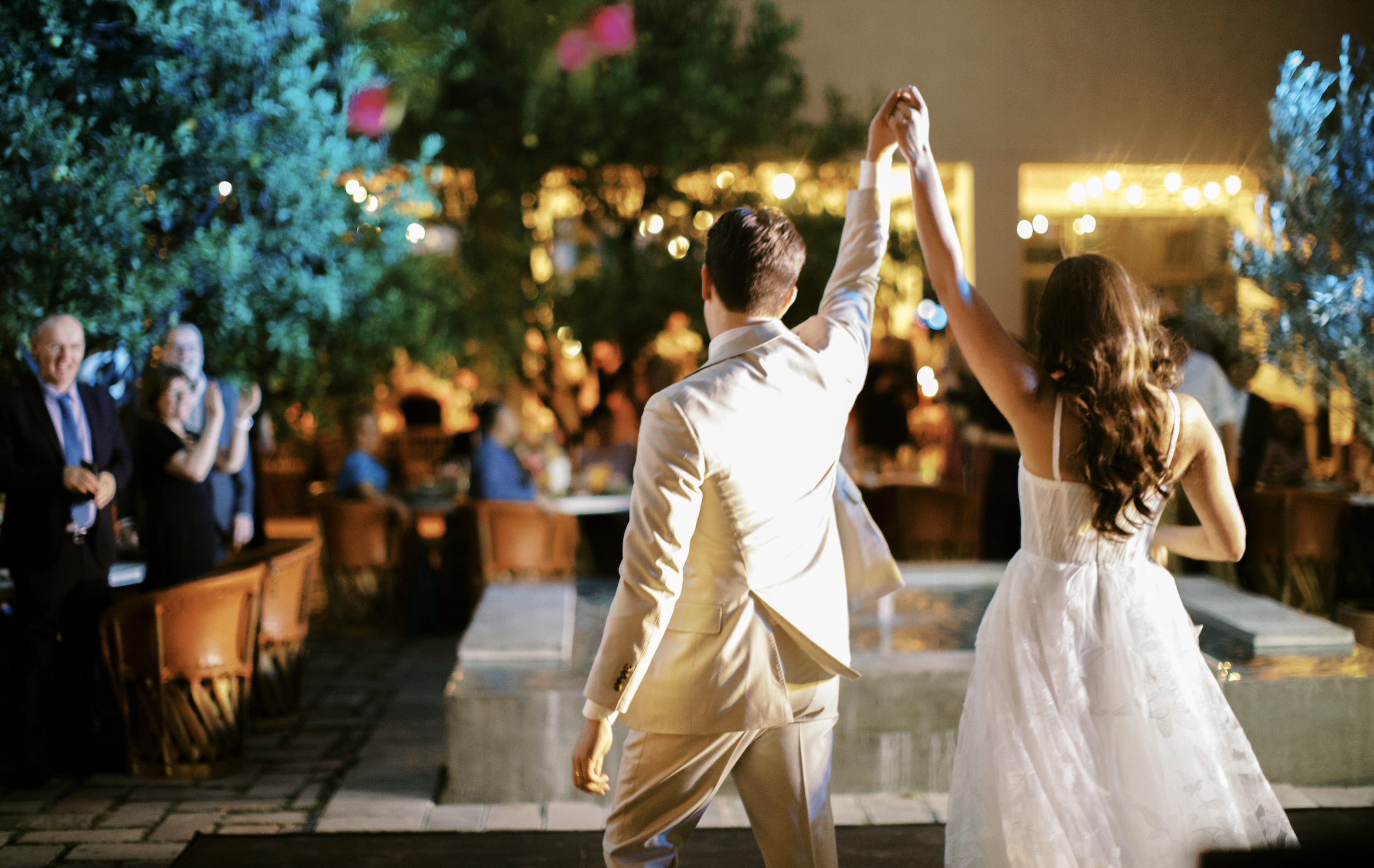 a bride and groom enter their dim lit wedding reception holding hands in the air as their guests clap 